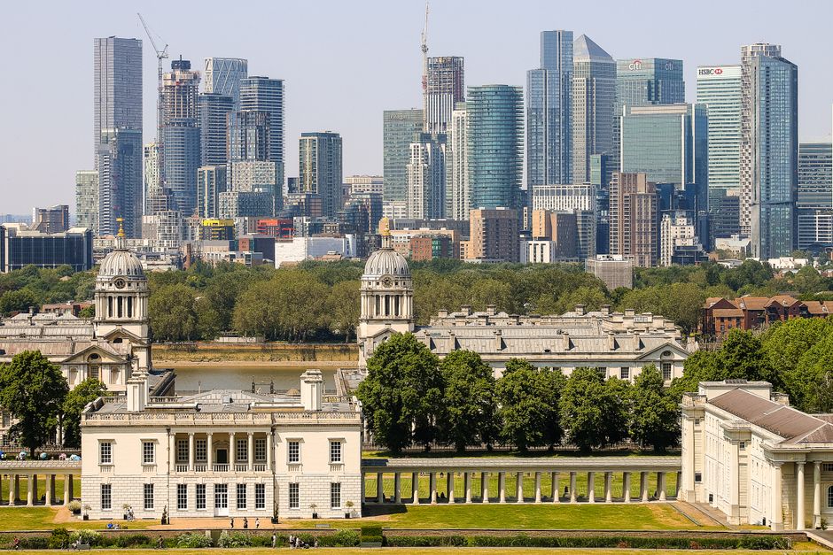 A wide cityscape view featuring a historic white building with classical architecture, including columns and domed towers, situated behind a row of lush green trees. In the background, there is a modern financial district with tall glass skyscrapers and high-rise office buildings, some under construction with cranes visible. The foreground includes a manicured lawn and a paved pathway along the historic building. The scene captures a contrast between the traditional architecture of the historic site and the contemporary skyscrapers of the city skyline, highlighting the area's blend of heritage and modern development. The image illustrates an urban environment suitable for house removals or furniture transport services, with the historic building indicating a distinguished local landmark often involved in relocation logistics managed by companies like Greenwich Removals, supporting professional packing and moving operations within the city.