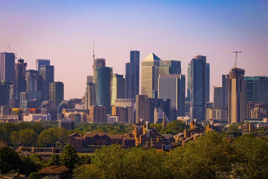 A city skyline featuring numerous modern high-rise office buildings and residential towers, with glass facades reflecting sunlight, set against a clear sky. In the foreground, there is a row of older, low-rise brick houses with pitched roofs and chimneys, bordered by lush green trees that add natural contrast to the urban environment. Visible construction cranes indicate ongoing development within the cityscape. The scene captures a moment during daylight hours in a bustling metropolitan area, representing a mix of contemporary and traditional architecture, which is relevant to the context of house removals and property relocation services offered by Greenwich Removals. The image emphasizes the urban environment where furniture transport, packing, and loading processes are undertaken as part of city-based moving and relocation logistics.