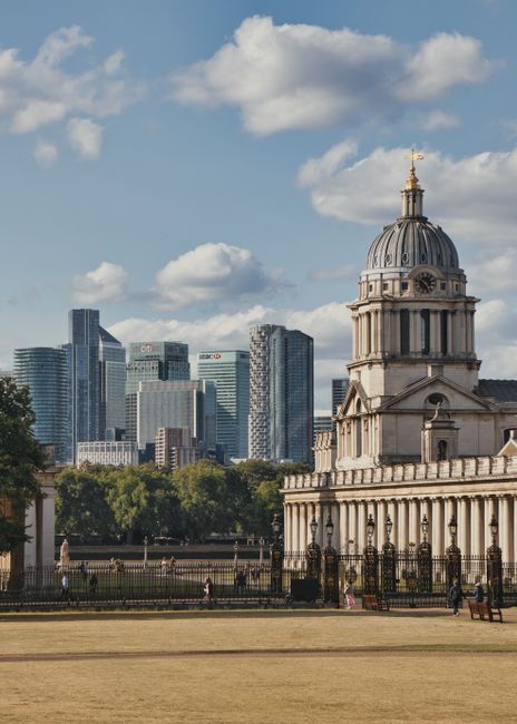 A wide cityscape view featuring a historic white building with classical architecture, including columns and domed towers, situated behind a row of lush green trees. In the background, there is a modern financial district with tall glass skyscrapers and high-rise office buildings, some under construction with cranes visible. The foreground includes a manicured lawn and a paved pathway along the historic building. The scene captures a contrast between the traditional architecture of the historic site and the contemporary skyscrapers of the city skyline, highlighting the area's blend of heritage and modern development. The image illustrates an urban environment suitable for house removals or furniture transport services, with the historic building indicating a distinguished local landmark often involved in relocation logistics managed by companies like Greenwich Removals, supporting professional packing and moving operations within the city.