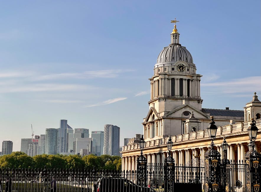 A large historic building with a prominent domed roof and clock face, surrounded by an ornate black wrought-iron fence with decorative lampposts. The structure features classical architectural details such as columns and arched windows. In the background, a modern city skyline with high-rise office buildings and cranes is visible against a clear blue sky, indicating an urban setting. The scene suggests a location in London, with the building potentially being a landmark associated with home relocation or furniture transport activities. The image depicts the exterior environment during daylight, emphasizing the contrast between historic architecture and modern urban development, relevant to the context of house removals and moving services provided by Greenwich Removals.