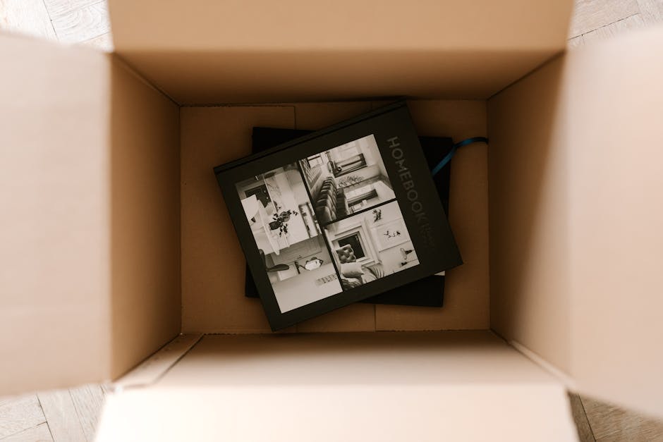 A cardboard moving box with flaps open, placed on a wooden floor, containing a black and white photo book titled 'Homebook' with images of furnished living rooms, sofas, and home interiors visible on the cover. The box appears ready for home relocation, with packing materials possibly supporting furniture transport and packing and moving activities. The interior environment is well-lit, and the image captures part of the loading process during house removals, as the box is positioned inside a property or on a pavement at the entrance, prepared for transport by Greenwich Removals as part of their removals service.