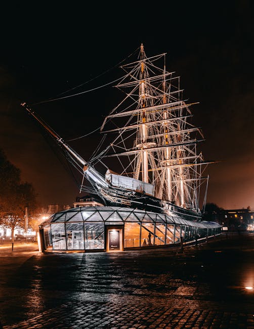 A large, historic sailing ship with multiple masts and rigging, illuminated against a dark night sky. The ship's hull and decks are visible, with some exhibit a metallic or wooden finish. It is situated on a paved area, possibly near or on the riverbank, with trees and distant city lights in the background. The ship appears to be part of a display or museum exhibit, enclosed by a glass structure at its base. The scene is well-lit, highlighting details of the ship's rigging and structure. This image, linked to Greenwich Removals, subtly reflects the planning and logistics involved in home relocation and furniture transport, such as careful packing, loading, and transportation of large objects, although the focus remains on the ship’s detailed presentation at night.