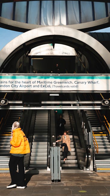 Inside a modern transportation hub featuring an entrance with a large circular archway and escalators leading to an upper level. The scene shows two people on the escalators, one woman wearing an orange jacket walking up and another person in a beige coat standing on the escalator, both engaged in their movements. The background displays signage related to Canary Wharf and transportation connections, with bright lighting illuminating the area. The space appears clean and well-maintained, with glass and metallic structural elements. This setting exemplifies the kinds of locations Greenwich Removals might coordinate for home relocation or furniture transport, especially when planning routes through busy urban areas like Greenwich. The image captures the logistical environment associated with packing, moving, and transport services involved in professional removals and relocation logistics.