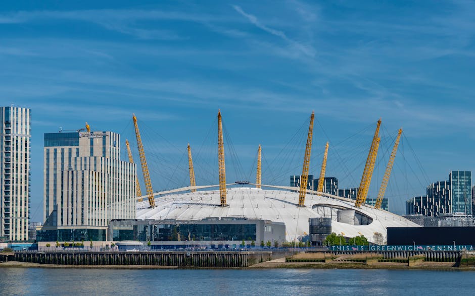 A view of the O2 Arena in North Greenwich, London, with its distinctive white, dome-shaped roof supported by numerous yellow masts and tension cables. To the left of the arena, several modern high-rise buildings with glass facades are visible, reflecting sunlight and contrasting with the arena's design. The scene is set along the river Thames, with a concrete and wooden jetty in the foreground. The sky above is partly cloudy, with streaks of blue and wispy clouds. Although the image primarily showcases the architectural landmark, it indirectly relates to house removals and furniture transport services, such as those provided by Greenwich Removals, especially for clients relocating in or near Greenwich or the O2 Arena area.