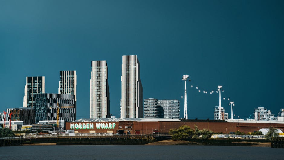 A wide-angle view of the North Greenwich waterfront showing modern high-rise office buildings and residential towers with reflective glass facades, situated behind Morden Wharf industrial building with a weathered brick exterior and a sign displaying 'Morden Wharf' in white and green letters. In the foreground, there are several large cardboard moving boxes, some wrapped in plastic and others stacked, positioned near the edge of the property. The boxes are being prepared for transportation as part of home relocation or furniture transport processes. A green lawn with a few small trees separates the boxes from a pathway or loading area where a moving van, branded with Greenwich Removals, is partially visible. The scene is illuminated by natural daylight with a partly cloudy sky, suggesting outdoor packing and loading activities related to house removals near the O2 Arena, Greenwich, reflecting professional moving logistics and loading operations.