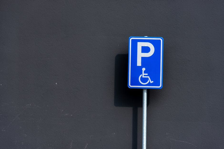 A blue accessibility parking sign with a white wheelchair symbol and a white letter 'P' mounted on a silver metal pole, positioned against a dark grey wall with a textured surface. The sign indicates
