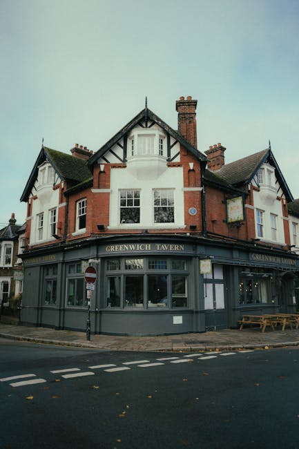 The exterior of Greenwich Tavern, a three-story building with a traditional brick and white facade, located at the corner of a street. The ground floor features large windows displaying interior lighting, with a sign reading 'Greenwich Tavern' above the windows, indicating it is a pub or restaurant. A small sidewalk surrounds the building, with a wooden bench positioned near the corner and a metal pole with traffic signs, including a no-entry sign, nearby. The upper floors have multiple sash windows, some with window boxes, and a steep, gabled roof with chimneys. The environment is a typical urban street scene, possibly during daylight hours with overcast skies. The image is relevant to relocation services and house moves, illustrating the type of property involved in home removals or furniture transport, as offered by Greenwich Removals.