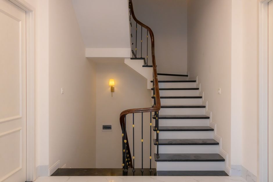 Photograph of a staircase inside a residential property, featuring white-painted walls and a curved wooden handrail with black metal balusters accented by gold-colored details. The stairs have black treads with white risers, leading up to an upper floor that is not visible in the image. To the left, part of a white doorframe is visible, while on the right, a section of plain wall is seen. A small, wall-mounted lamp with a yellow shade provides ambient lighting, located near the corner of the staircase. The environment appears clean and well-maintained, with a smooth, polished floor surface at the base of the stairs. The scene depicts an interior space that may present access challenges for furniture or boxes during a home relocation, and highlighting the importance of planning for stair access issues, which Greenwich Removals can assist with during packing and moving projects in Greenwich.