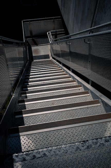 The image shows a set of external metal stairs with diamond plate treads leading downward, accompanied by metal railings on both sides. The stairs are situated on a building's exterior, with a dark overhang above providing some shade. The staircase is positioned adjacent to a wall and appears to be part of an emergency exit or access route for a residential or commercial property. The lighting suggests daylight conditions, with shadows cast across the steps. This type of stair access can present challenges for furniture transport during home relocation, especially when tight spaces or narrow doorways are involved, making proper planning essential. Greenwich Removals highlights such considerations in their tips for managing stair access issues during house moves, emphasizing the importance of careful handling and possibly specialized equipment to facilitate safe and efficient furniture transport in confined areas.